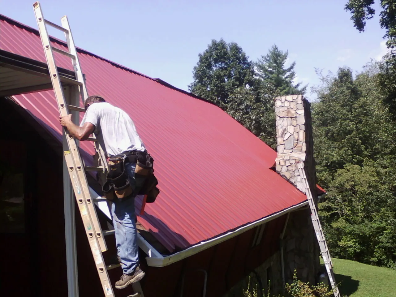 Roofer on a ladder beside a red metal roof and stone chimney — Soffit & Fascia Repair by Roofing Brothers in Conneautville